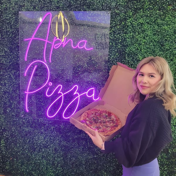 Woman holding an Apna Pizza box with a pizza, standing by a neon sign on a green wall.