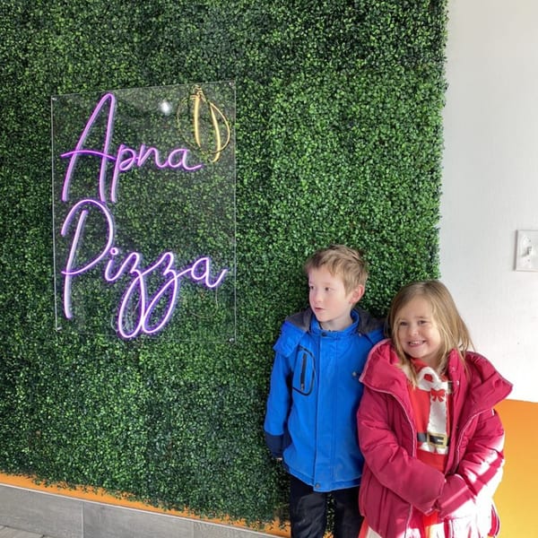Two children stand in front of a green hedge wall with a glowing purple "Apna Pizza" neon sign.