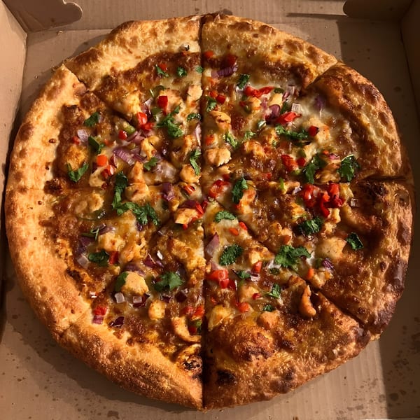 Apna Pizza's chicken tikka pizza, topped with red onions, bell peppers, and cilantro, in a delivery box.