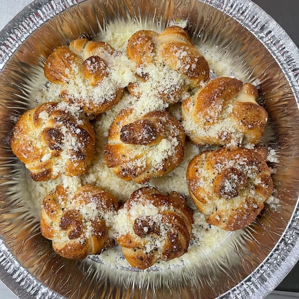 Seven golden-brown garlic knots, generously topped with grated Parmesan cheese, in a foil pan.