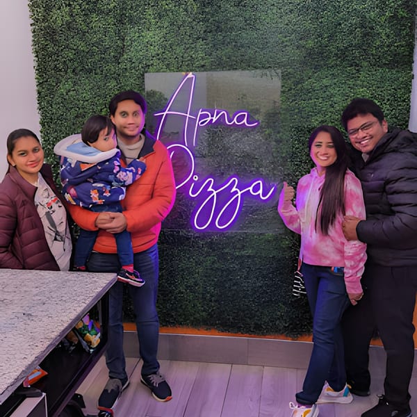 A family of five, including a baby, smiling in front of a neon "Apna Pizza" sign.
