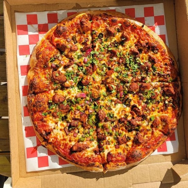 A whole Apna Pizza, topped with chicken, green peppers, and cilantro, in a delivery box.